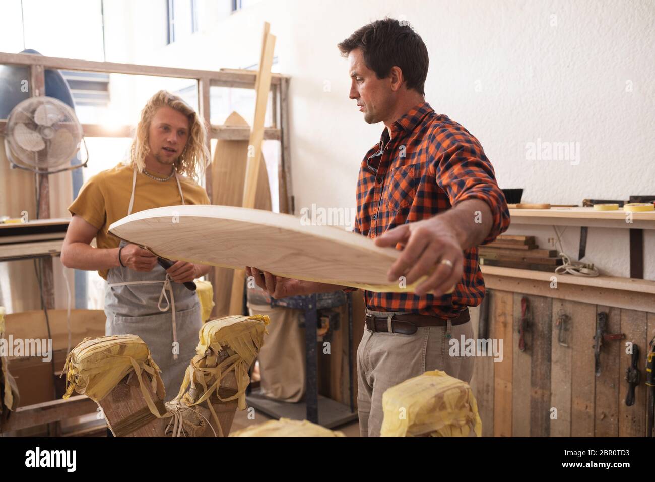Due produttori caucasici di tavole da surf maschili che fanno insieme una tavola da surf in legno e la ispezionano Foto Stock