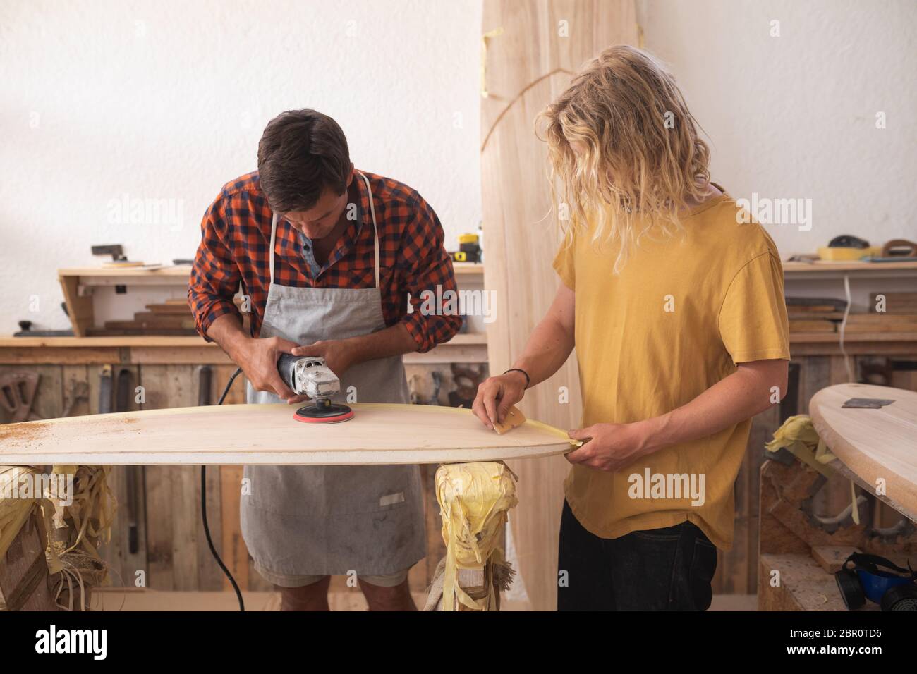 Due produttori caucasici di tavole da surf maschili che fanno una tavola da surf in legno insieme ad una levigatrice Foto Stock