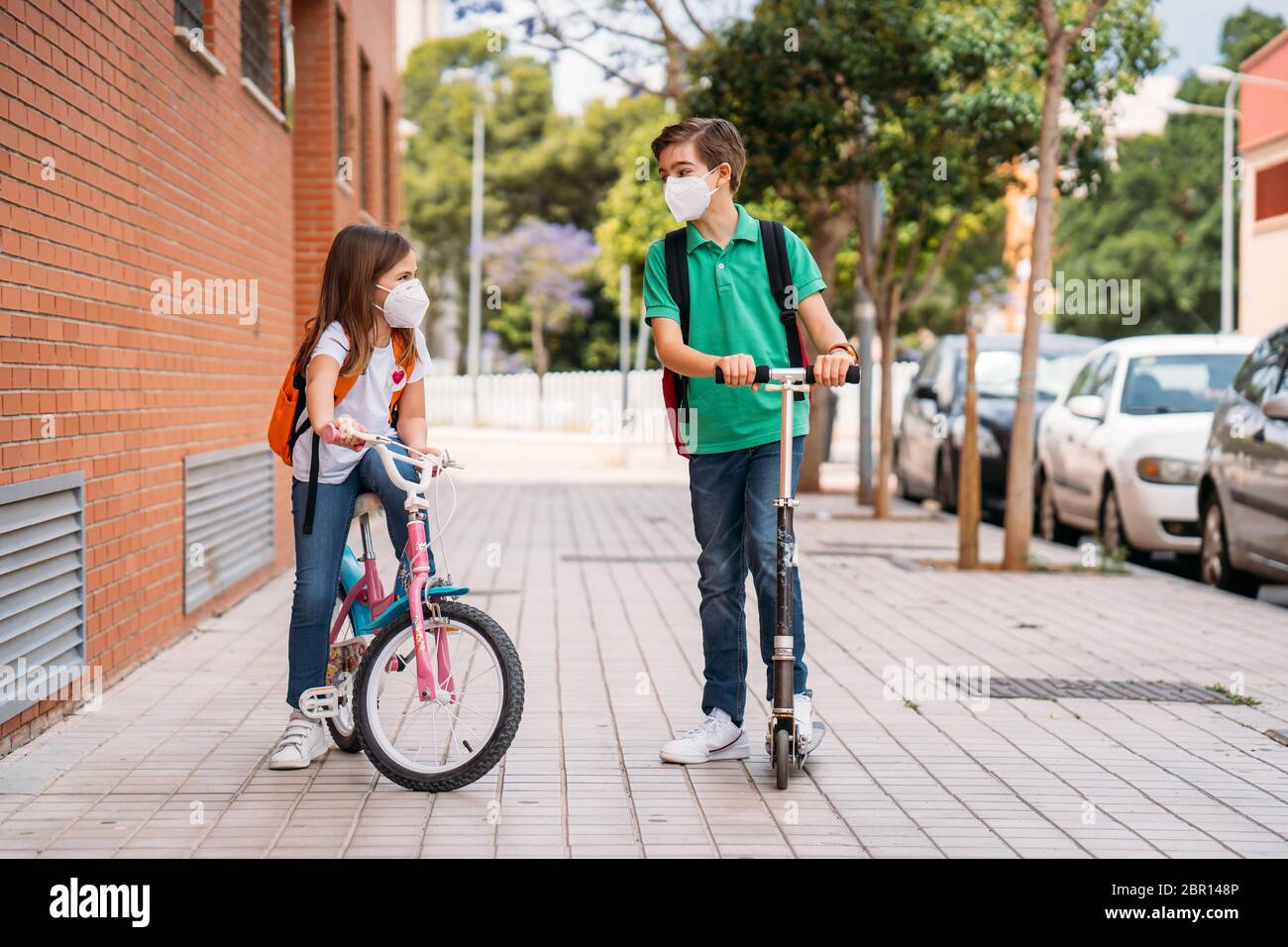 Ragazzo e ragazza che indossano maschere e cavalcano uno scooter e una bicicletta sulla strada Foto Stock