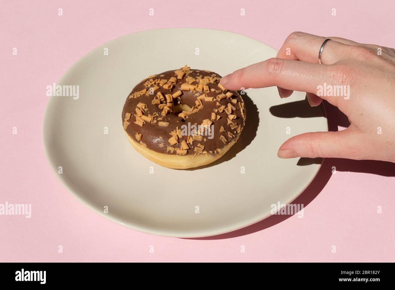 Una mano con le frettole sta prendendo donat cioccolato decorato con ricci al caramello sulla parte superiore al piatto bianco sullo sfondo rosa chiaro e soleggiato Foto Stock