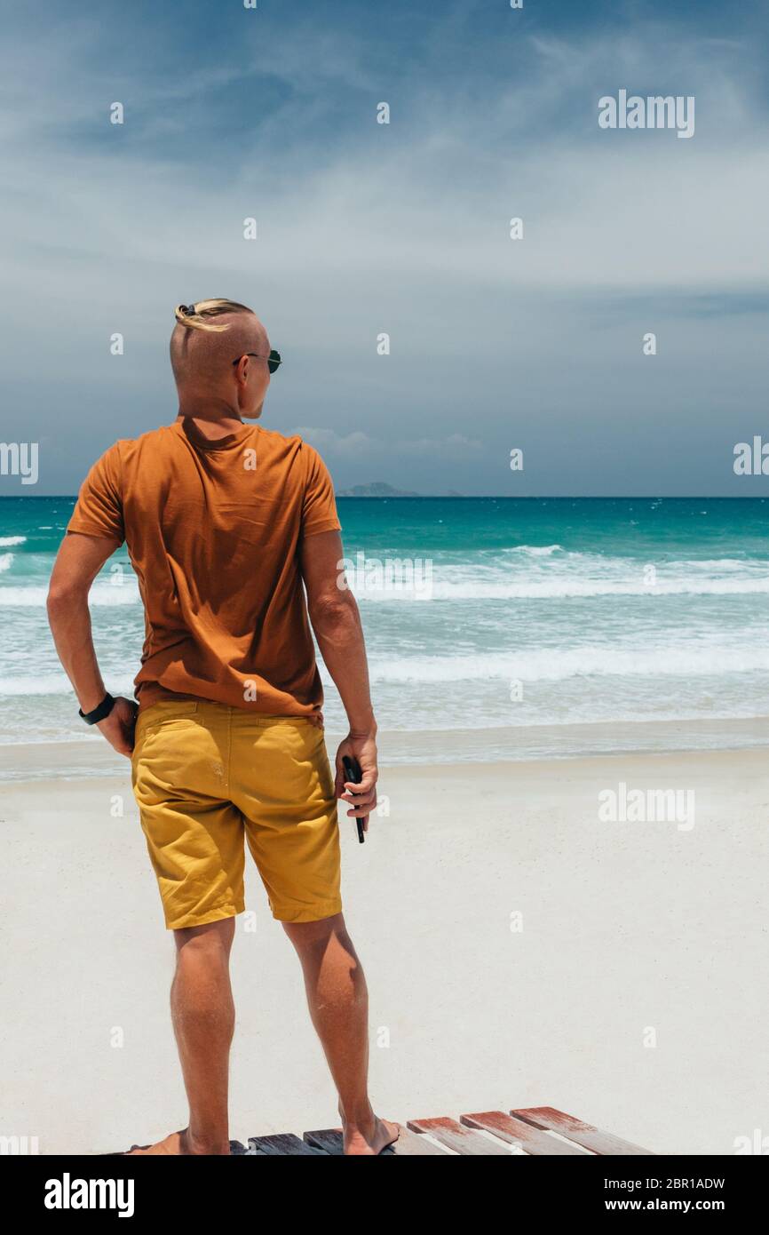 Giovane ragazzo in una T-shirt marrone e pantaloncini in piedi sulla spiaggia sabbiosa in occhiali da sole. Giorno estivo chiaro, cielo con nuvole bianche, onde con f bianco Foto Stock