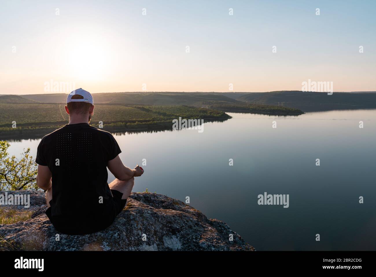Concetto di meditazione yoga benessere. Giovane uomo seduto alla macchina fotografica in posizione lotus sulla roccia sotto il fiume tropicale Foto Stock