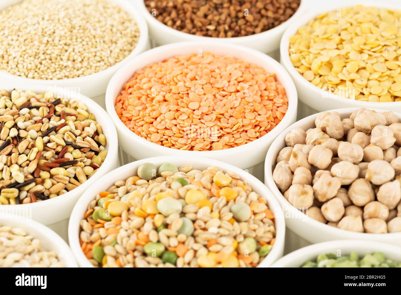 La collezione di diversi tipi di groat nelle ciotole in ceramica bianca. Piselli spezzati, lenticchie colorate, grano saraceno, ceci, grani di grano, quinoa. Foto Stock