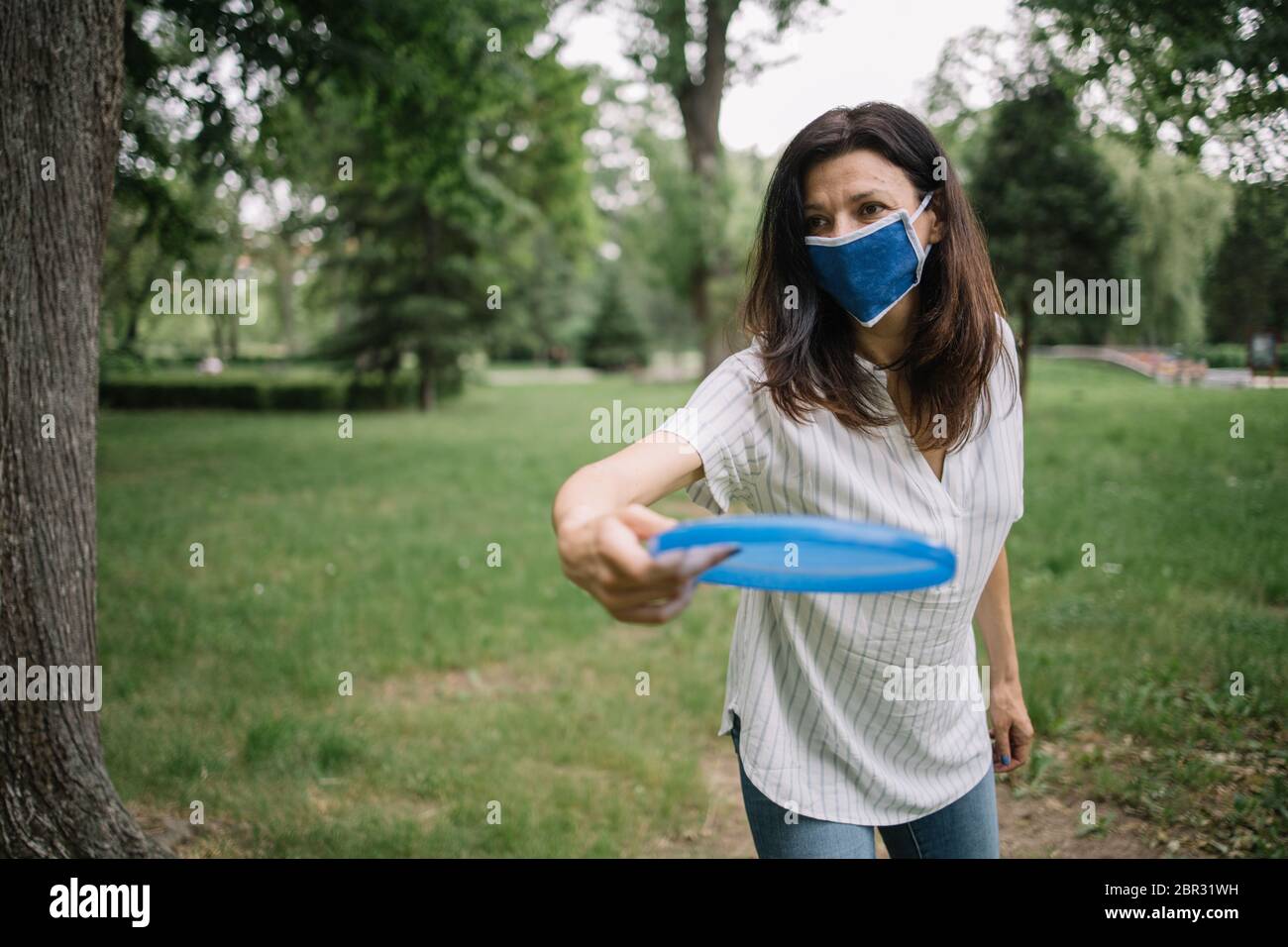 Giovane donna con maschera medica che getta disco volante. Donna di brunette che gioca frisbee nel parco dopo l'allentamento delle restrizioni del coronavirus Foto Stock