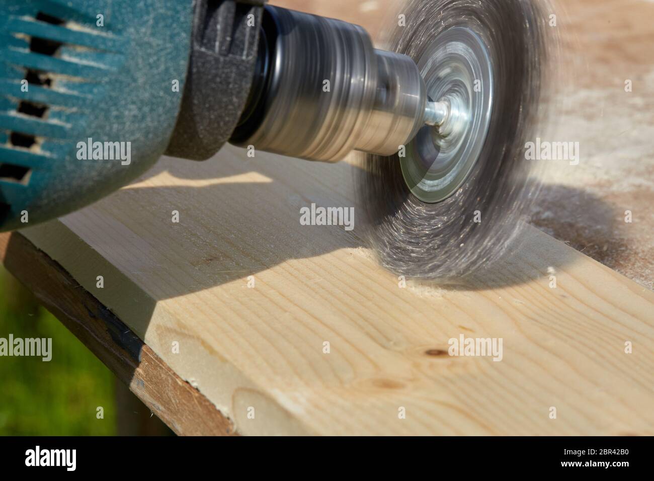 Il processo di spazzolatura di tavola di legno. La mano maschio sta tenendo la spazzolatrice elettrica che ruota con il disco metallico della spazzola che leviga un pezzo di legno. Foto Stock