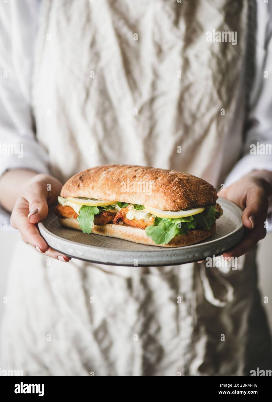 Donna in grembiule di lino con panino di pesce fresco fritto con salsa tartare, limone e rucola, fuoco selettivo. Idee per la colazione salutari e facili Foto Stock