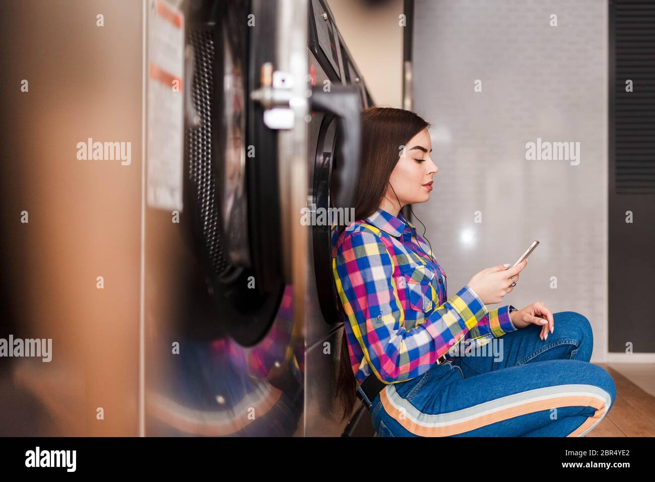 Ragazza seduta sul pavimento in una lavanderia e ascoltando musica con le cuffie. Concetto di processo di lavanderia Foto Stock