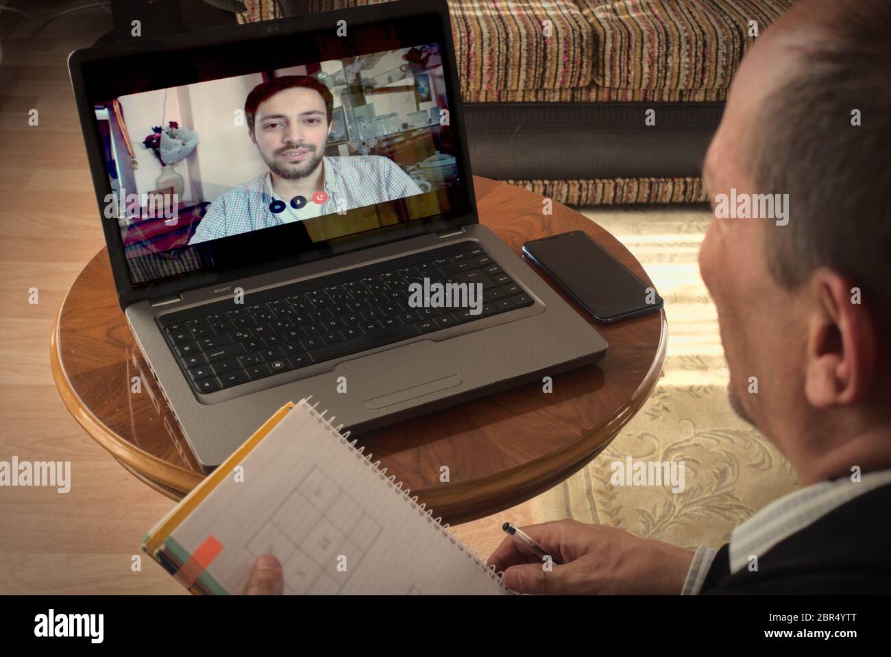 Un esperto che lavora da casa con il suo laptop, parla video con il suo collega più giovane e prende appunti Foto Stock