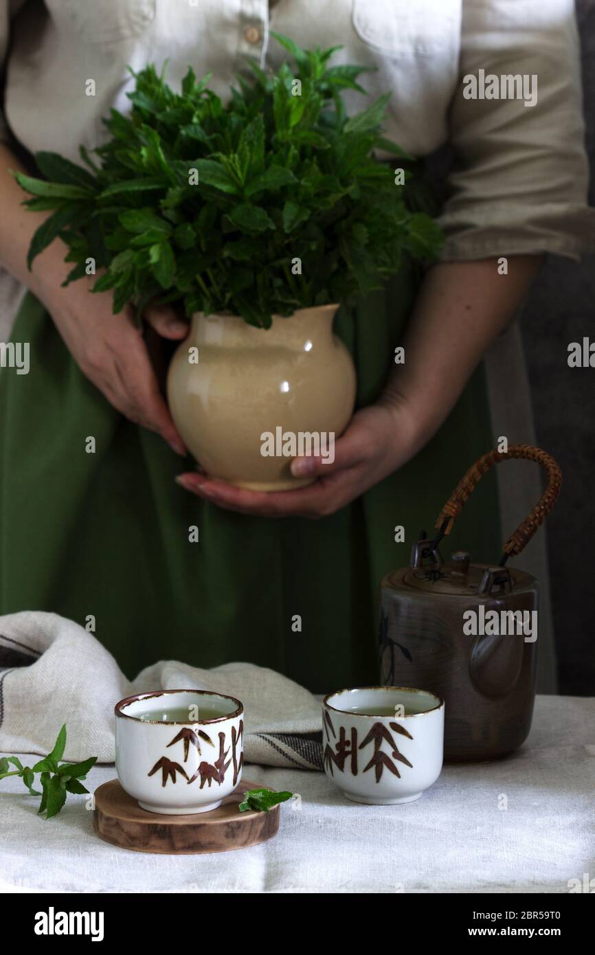 Tè alla menta in un set da tè cinese d'epoca, una donna in un abito di lino e un grembiule, e un bouquet di menta in una caraffa. Foto Stock