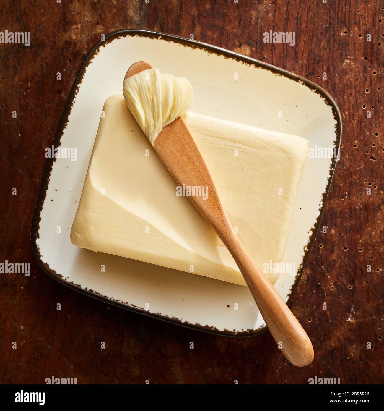 Pat di burro di fattoria con diffusore in legno con un cucchiaio di burro sulla parte superiore visto dal di sopra su una tavola in legno rustico in formato quadrato Foto Stock