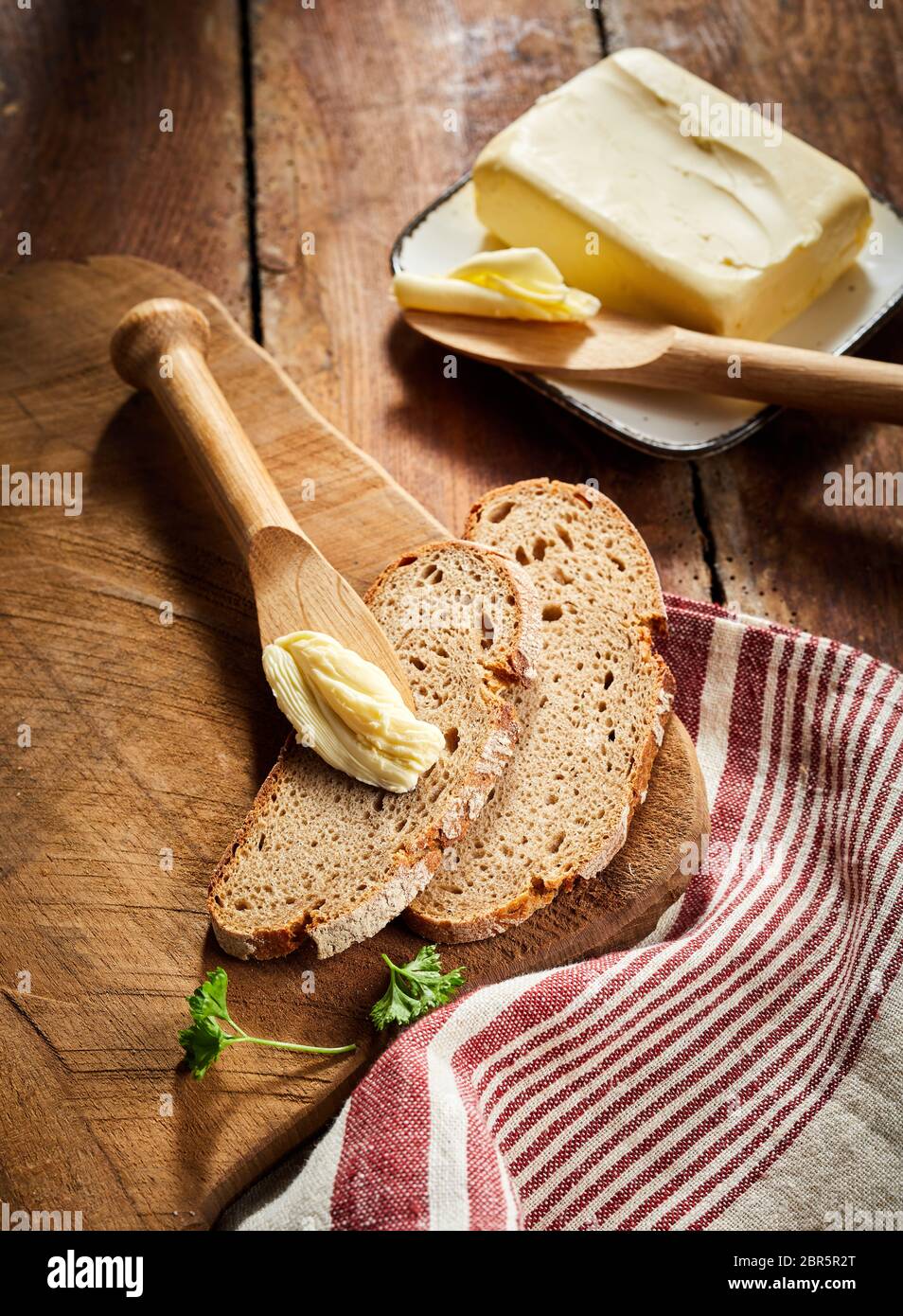 Pat di burro di fattoria con fette di pane di segale e un diffusore in legno o il coltello su un tagliere di legno con striping panno rustico Foto Stock