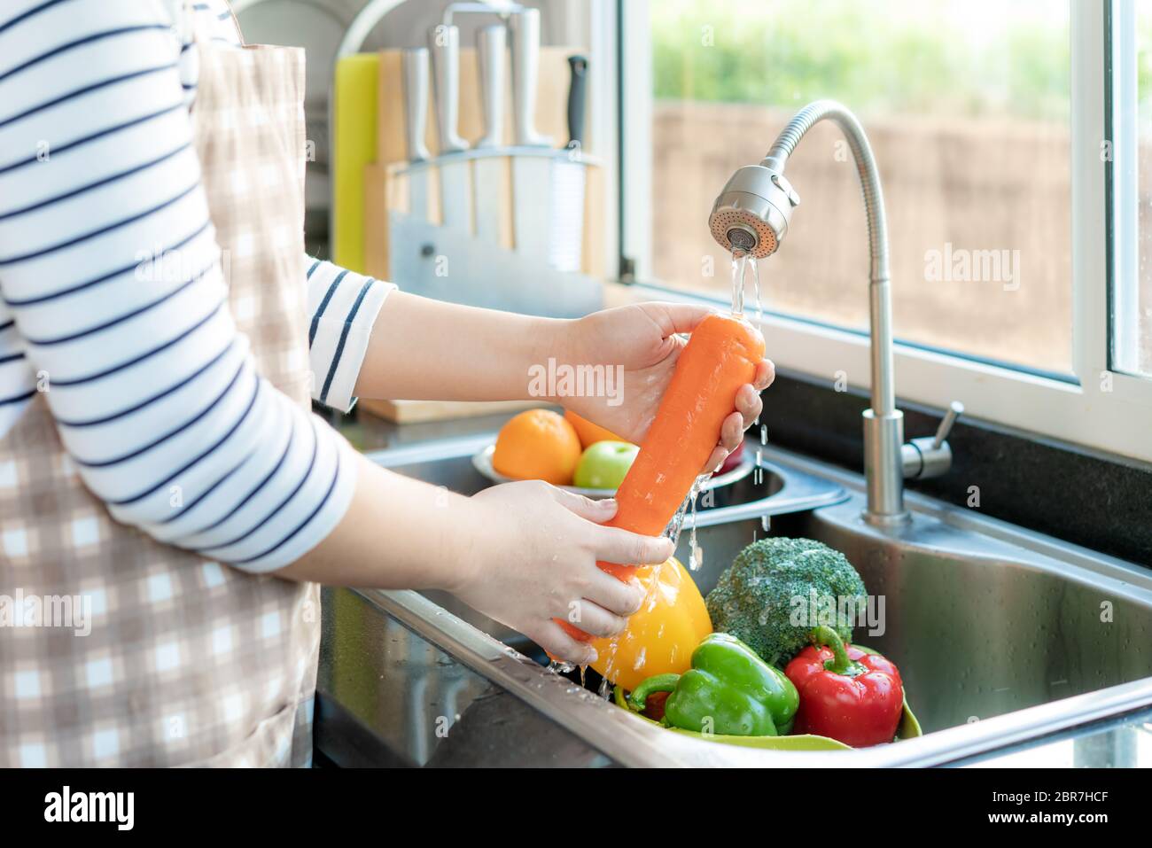 Asian donna sana lavando una carota e altre verdure sopra lavello della cucina e pulendo una frutta / verdura con acqua per eliminare le possibilità di c Foto Stock