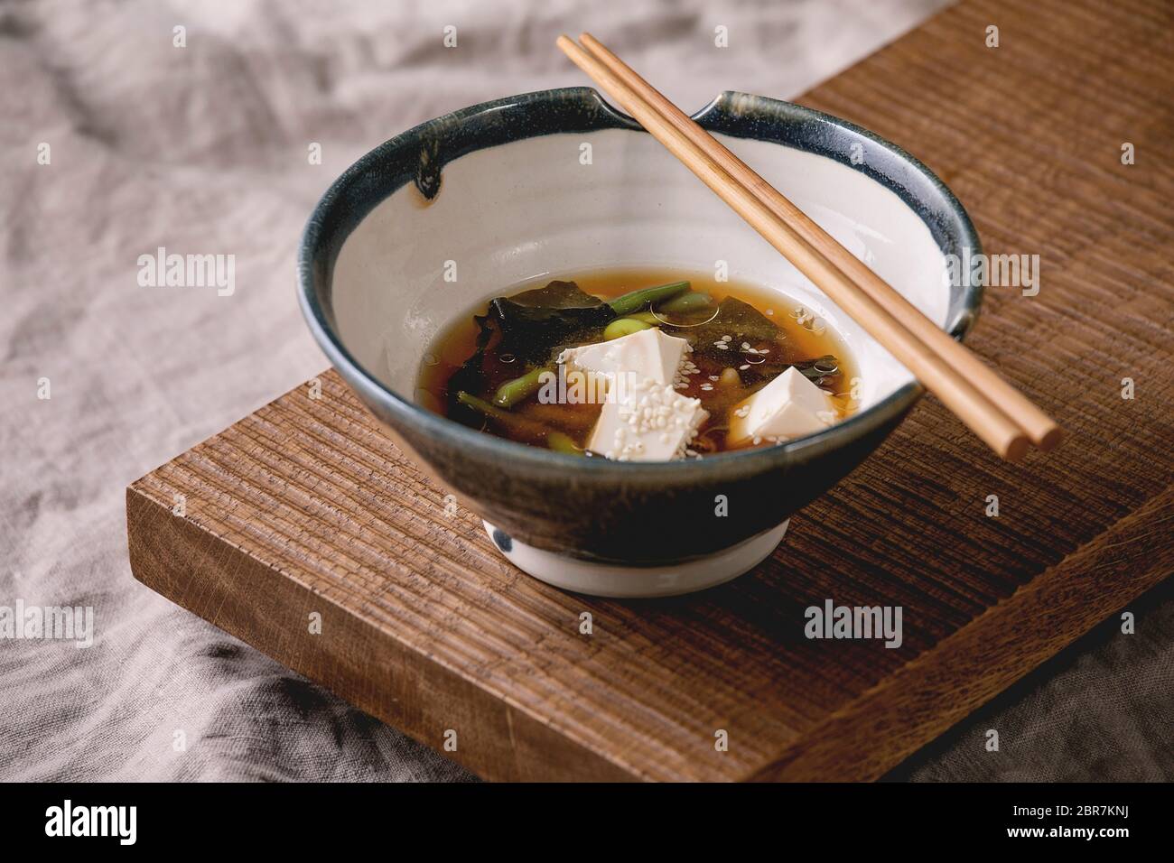 Zuppa di brodo misto giapponese con cubetti di tofu di seta, edamame di soia, fagioli verdi in ciotola tradizionale con bacchette su tavolo giapponese di legno su lin grigio Foto Stock