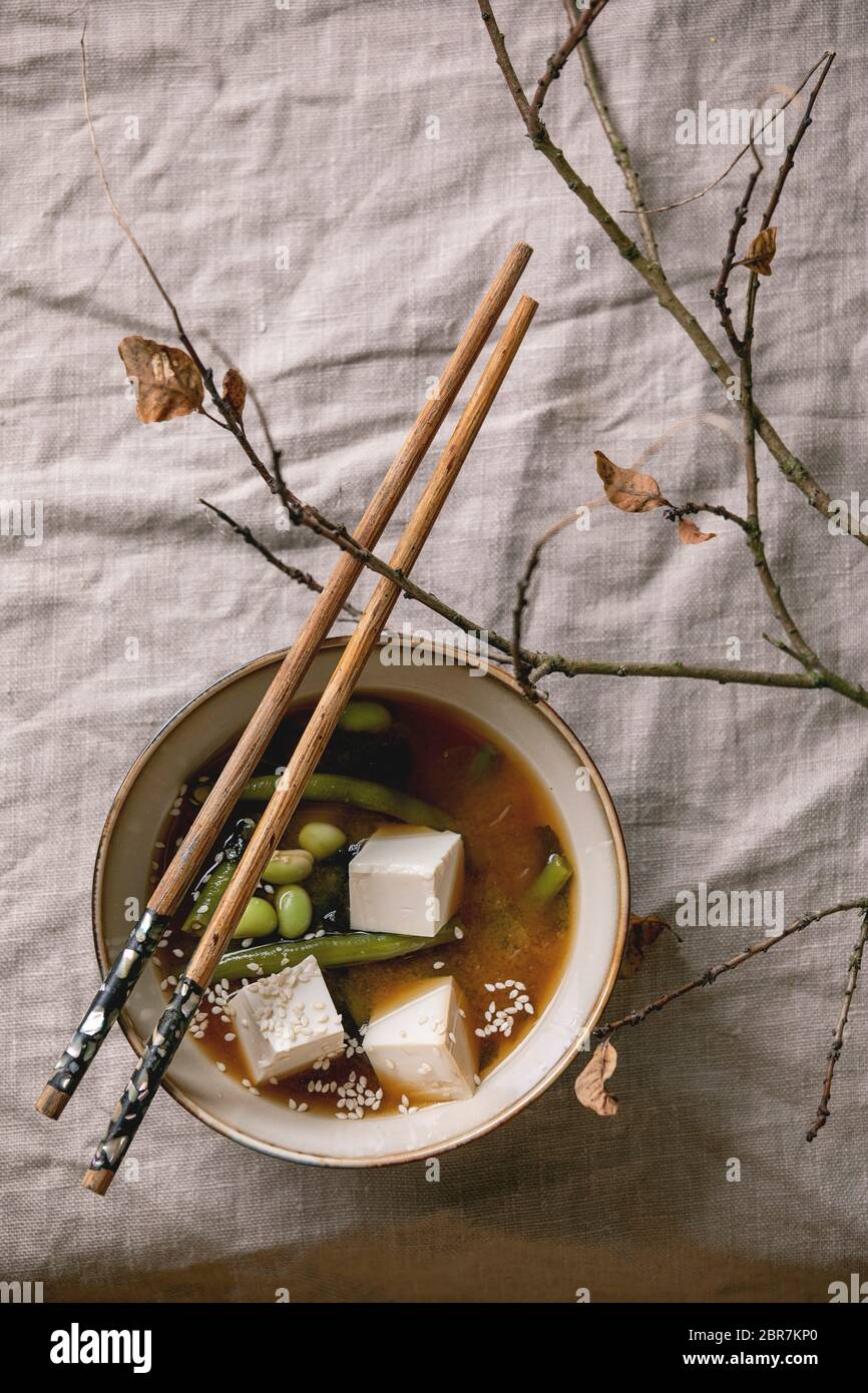 Zuppa giapponese di brodo miso con cubetti di tofu di seta, edamame di fagioli di soia, fagioli verdi in ciotola di ceramica con bastoncini di cotone su tovaglia di lino grigio con br autunno Foto Stock