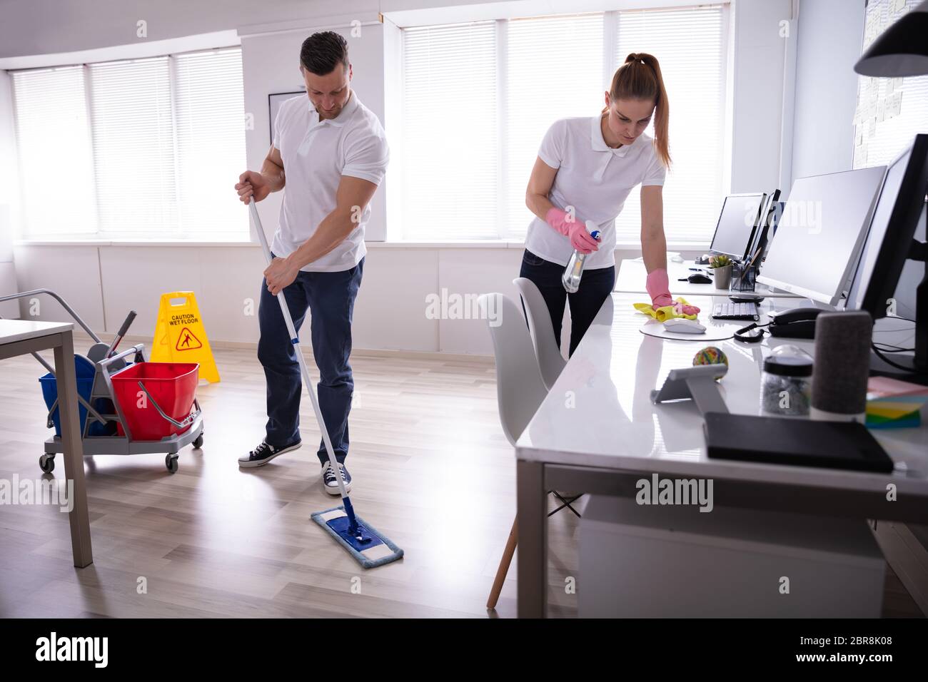Due giovani sorridenti bidello di pulizia della scrivania e pavimento con straccio In ufficio Foto Stock