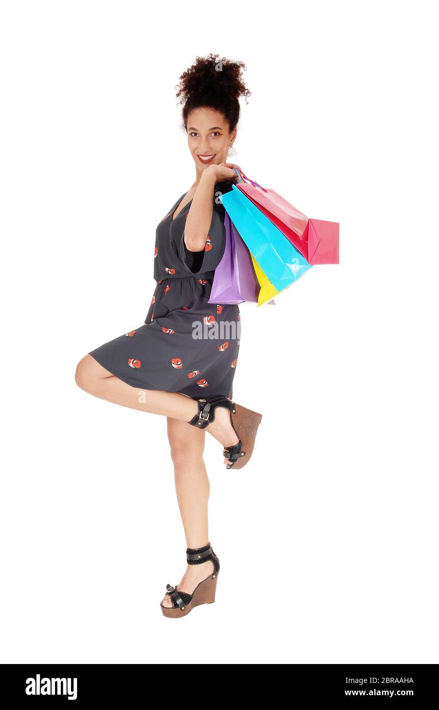 Un felice multi-razziale donna in piedi su una gamba sola tenendo le sue borse per lo shopping sulla sua spalla sorridente nella fotocamera con la sua parentesi capelli neri, isolato Foto Stock