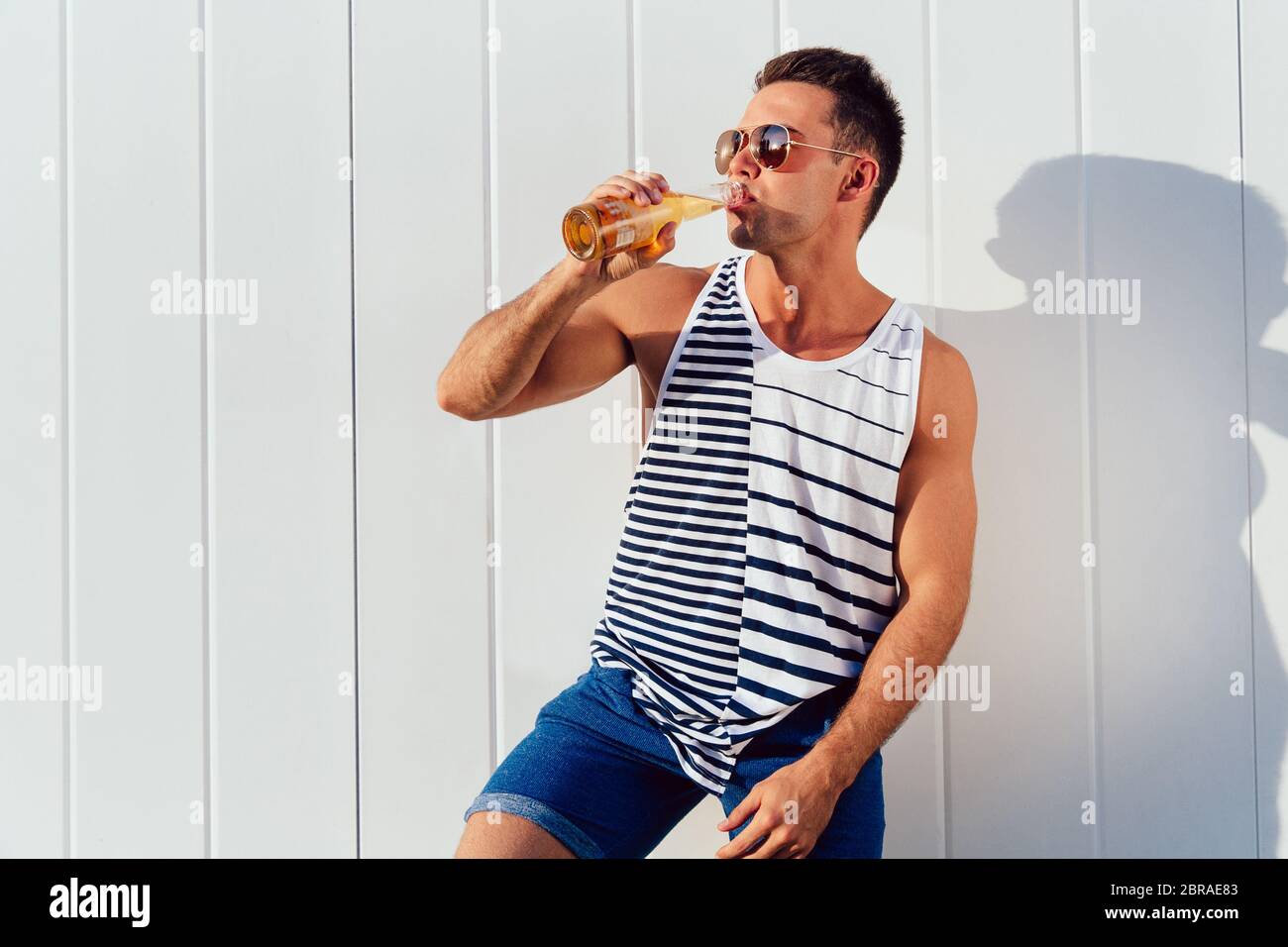 Attraente muscolare uomo in occhiali da sole di bere birra fredda, godendo le calde giornate estive, all'esterno. Vestito in un elegante canotta e shorts. Profilo. Foto Stock