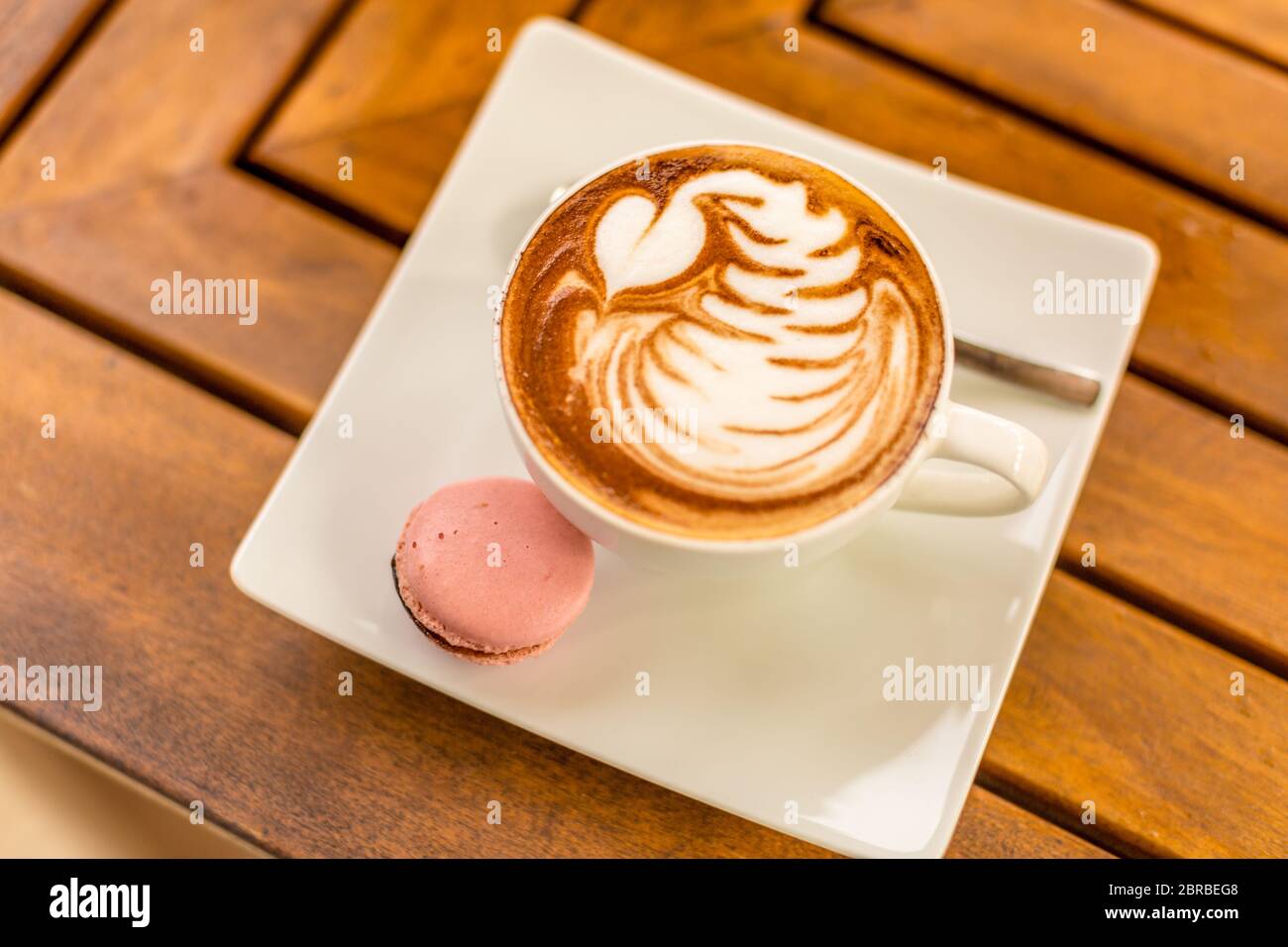 Primo piano di cappuccino latte caldo con bella latte art in una tazza di ceramica bianca su uno sfondo di tavola di legno e biscotti macaron Foto Stock