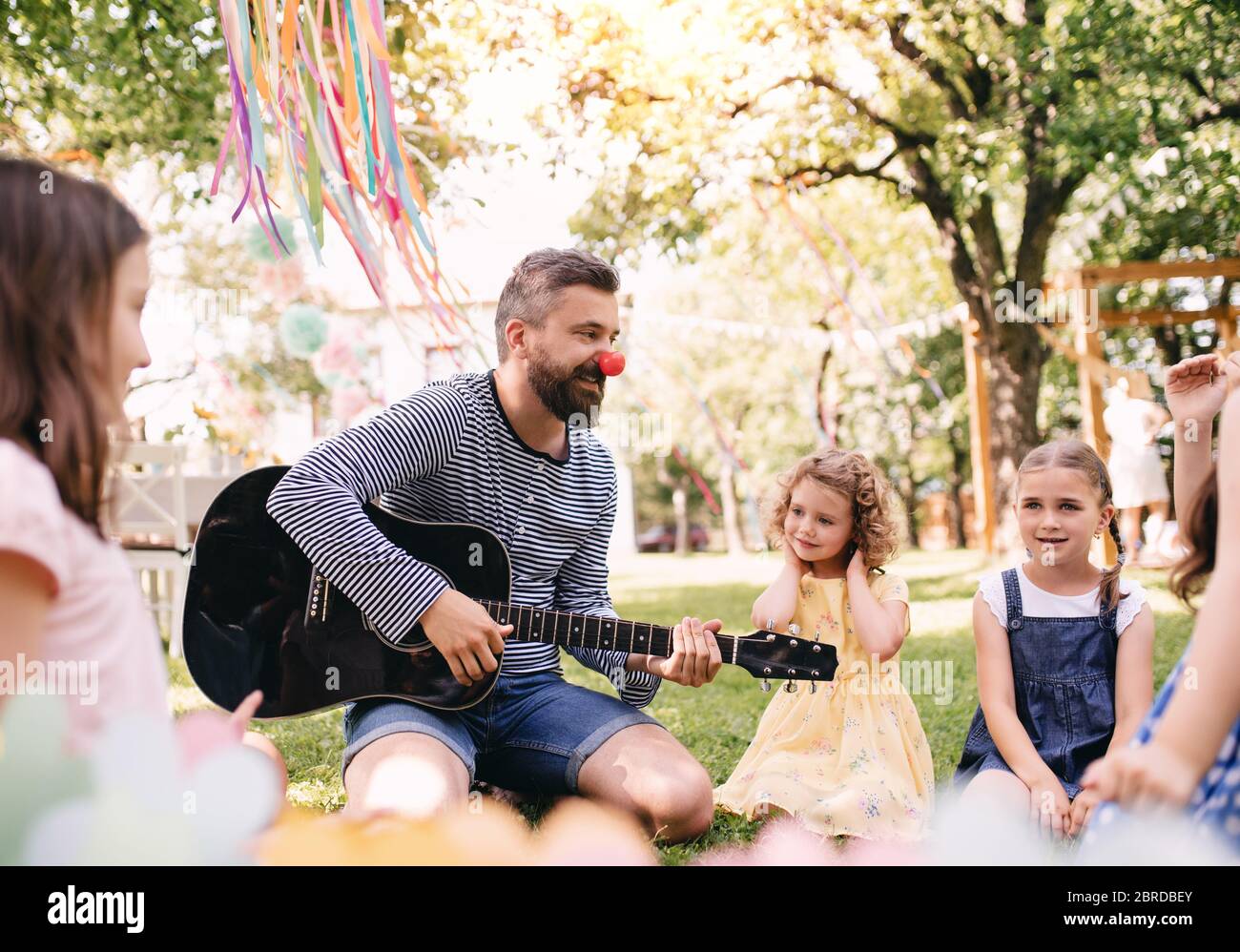 Uomo con bambini piccoli a terra all'aperto in giardino in estate, suonando la chitarra. Foto Stock