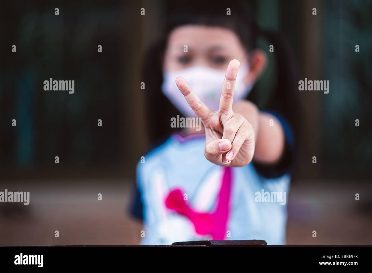 Asian bambina closeup ragazza sollevare due forma V dito indossare una maschera per la sicurezza Coronavirus per sostenere nella lotta contro l'epidemia di malattia COVID 19 c Foto Stock