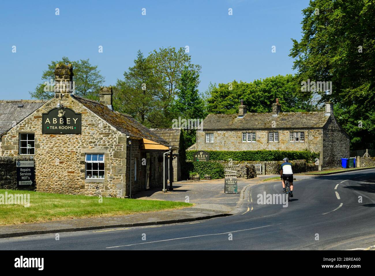 1 cyclist in bicicletta, pedalando su strada di campagna passando davanti al pittoresco caffè delle sale da tè in un pittoresco villaggio rurale soleggiato - Bolton Abbey, North Yorkshire, Inghilterra, Regno Unito Foto Stock