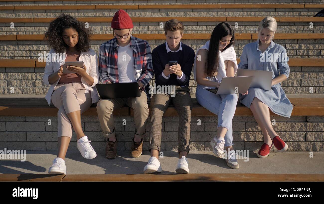 Studenti multietnici seduti in fila assorbiti da telefoni, compu Foto Stock