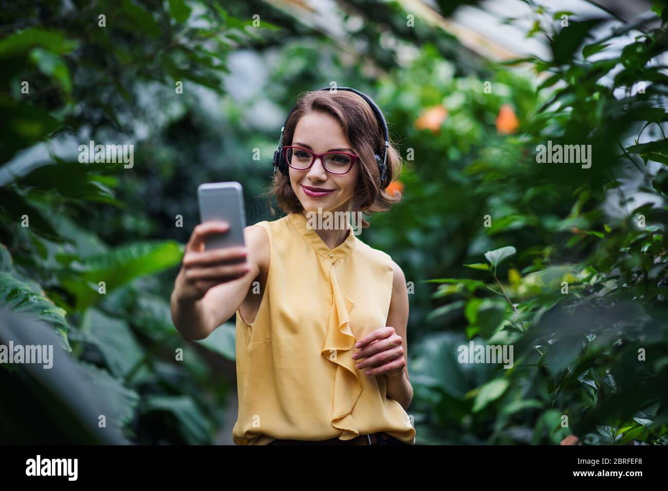 Giovane donna con smartphone in piedi in giardino botanico, prendendo selfie. Foto Stock