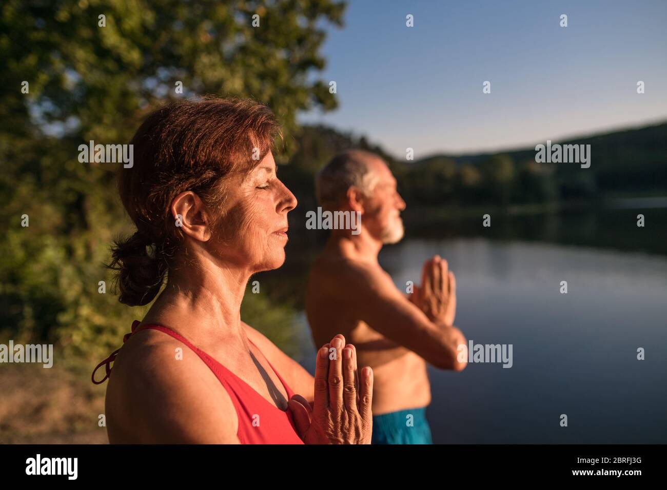 Coppia senior in costume da bagno in piedi vicino al lago all'aperto facendo yoga. Foto Stock