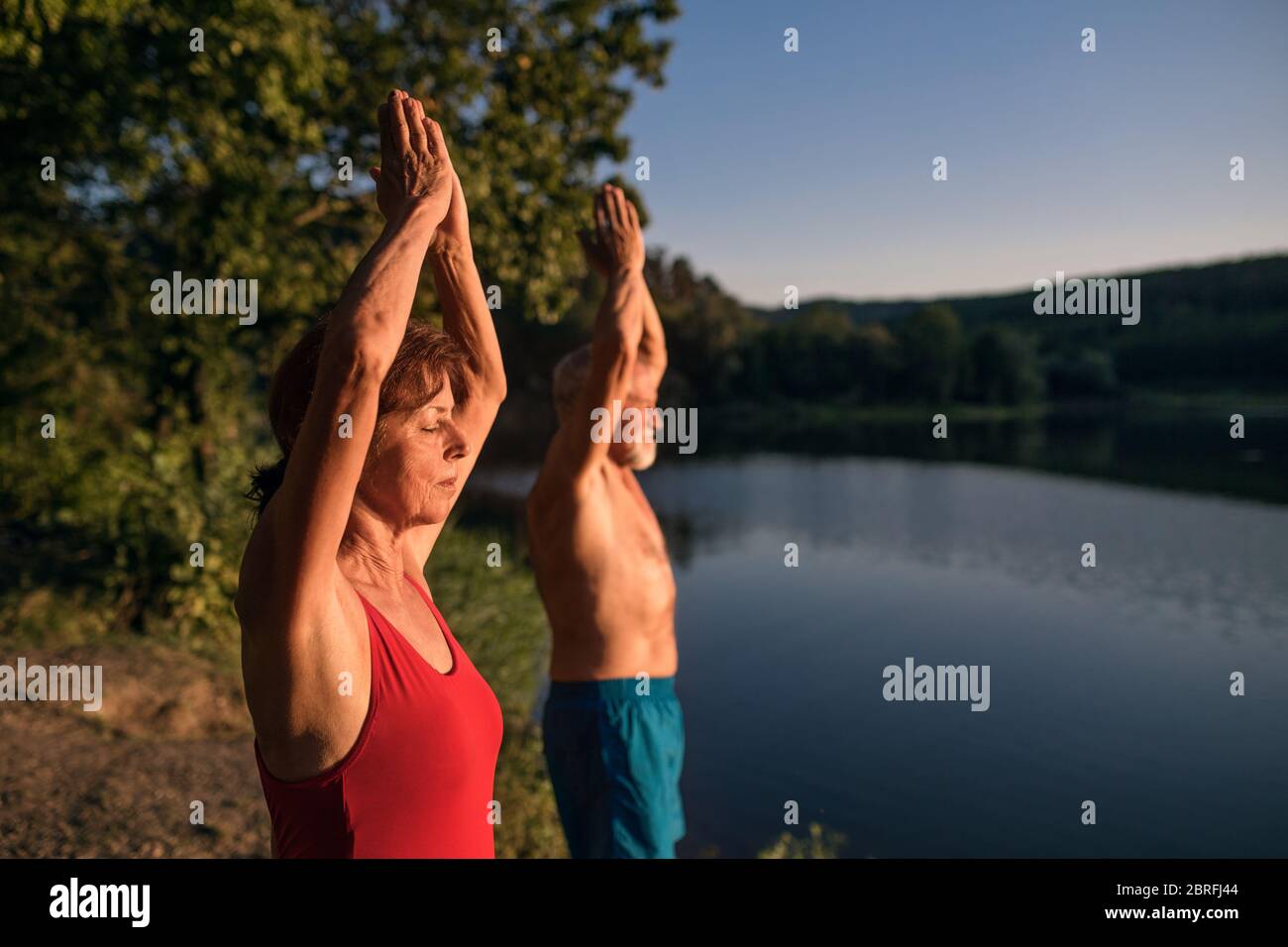 Coppia senior in costume da bagno in piedi vicino al lago all'aperto facendo yoga. Foto Stock
