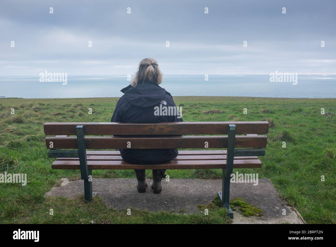 Una donna solita si siede su una panchina del parco vicino alla costa che guarda al mare in lontananza Foto Stock
