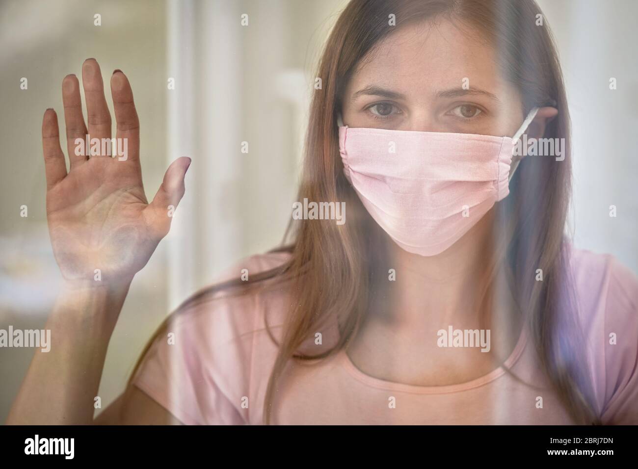 Giovane donna in rosa cotone viso virus maschera in piedi dietro vetro finestra vetro pannello, toccandolo con la mano, guardando triste. Quarantena o soggiorno a casa per essere saf Foto Stock