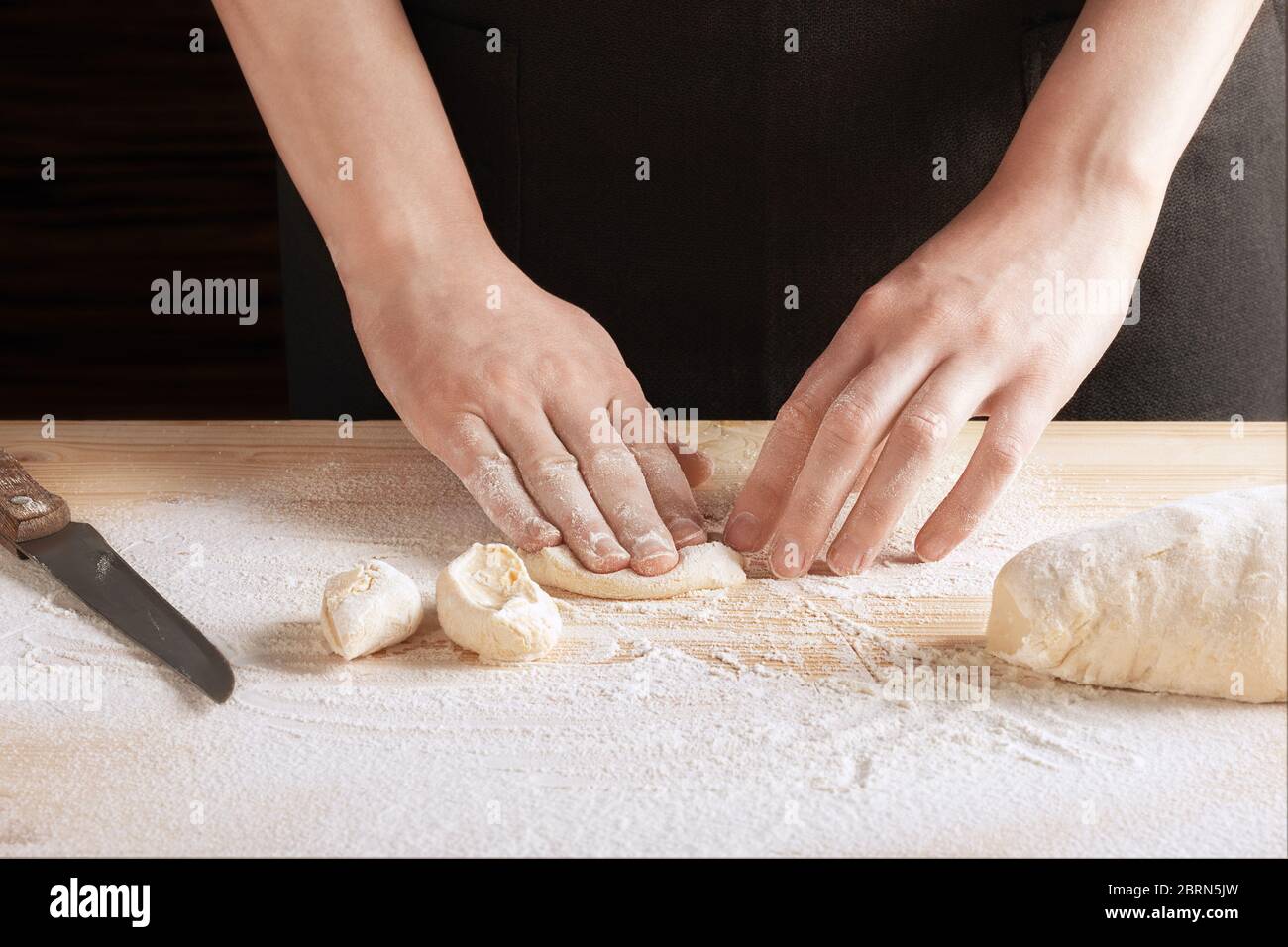 Vista frontale delle mani femminili che impastano una fetta di impasto su un tavolo di legno cosparso di farina. Una ricetta passo-passo per cucinare ravioli o gnocchi, s Foto Stock