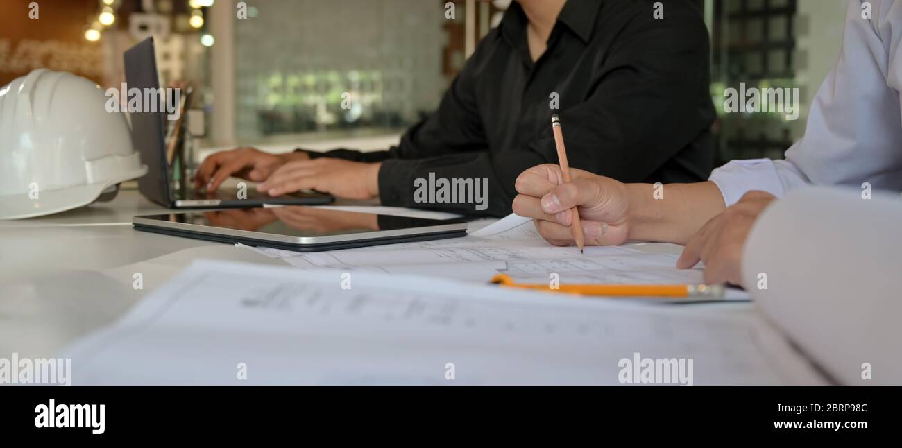 Un'immagine ritagliata di due ingegneri che lavorano insieme a BluePrint, tablet digitale e notebook sulla scrivania dell'ufficio Foto Stock