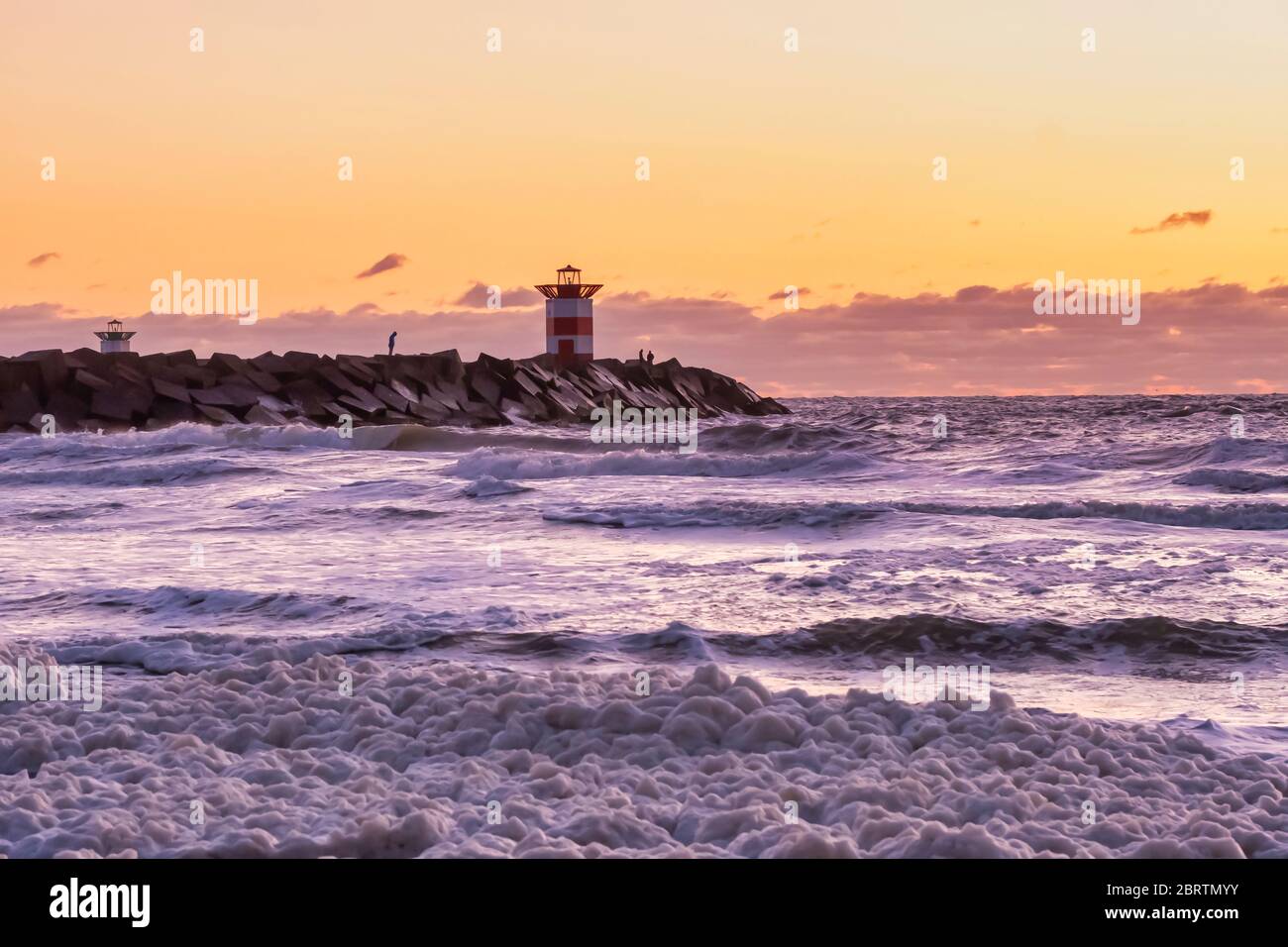 Faro a strisce rosse e bianche durante un tramonto vibrante. L'acqua colpisce i massi neri nelle onde. Cielo spettacolare in colore dorato. Scheveningen, il Neth Foto Stock