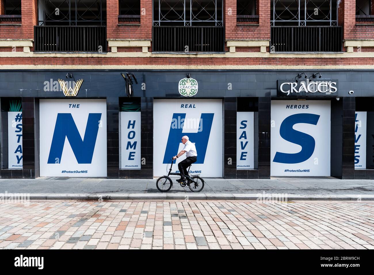 Coronavirus lockdown - un vecchio ciclista maschile passa un gigante 'NHS Stay Home Save Lives' segno nelle finestre del pub chiuso - Glasgow, Scozia, UK Foto Stock
