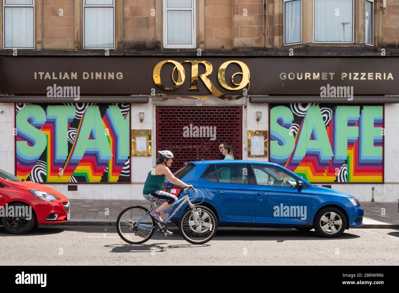 Donna che cavalcava una bicicletta durante il blocco pandemico del coronavirus, passato un murale 'sicuro' a Shawlands, Glasgow, Scozia, Regno Unito Foto Stock