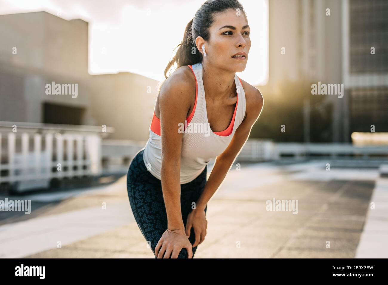 Atleta femminile che si appoggia in avanti con le mani sulle ginocchia e si guarda via. Donna runner che si prende una pausa dopo la corsa mattutina. Foto Stock