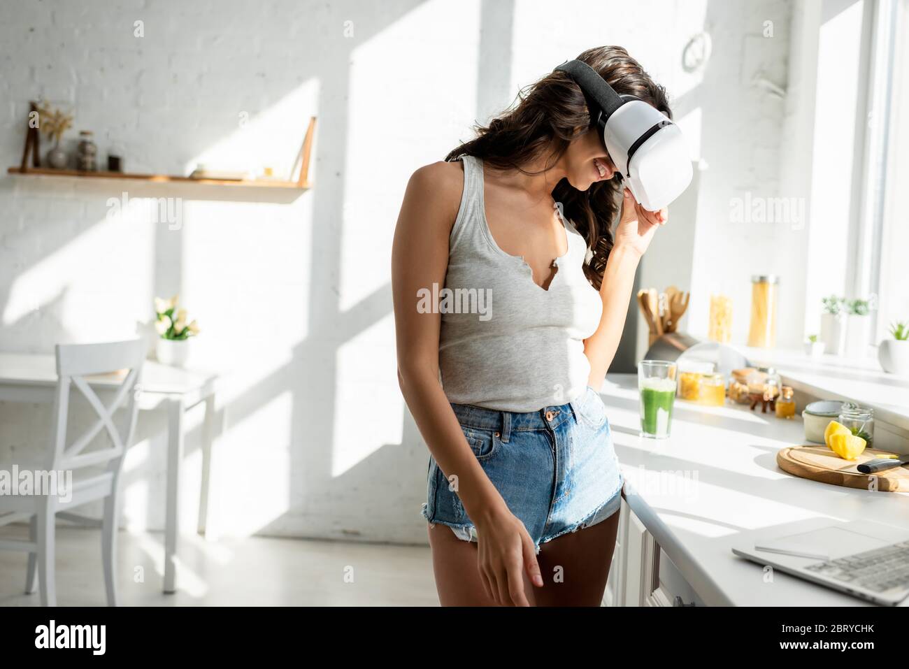 Messa a fuoco selettiva di donna sorridente in una cuffia per realtà virtuale in piedi vicino al laptop in cucina Foto Stock