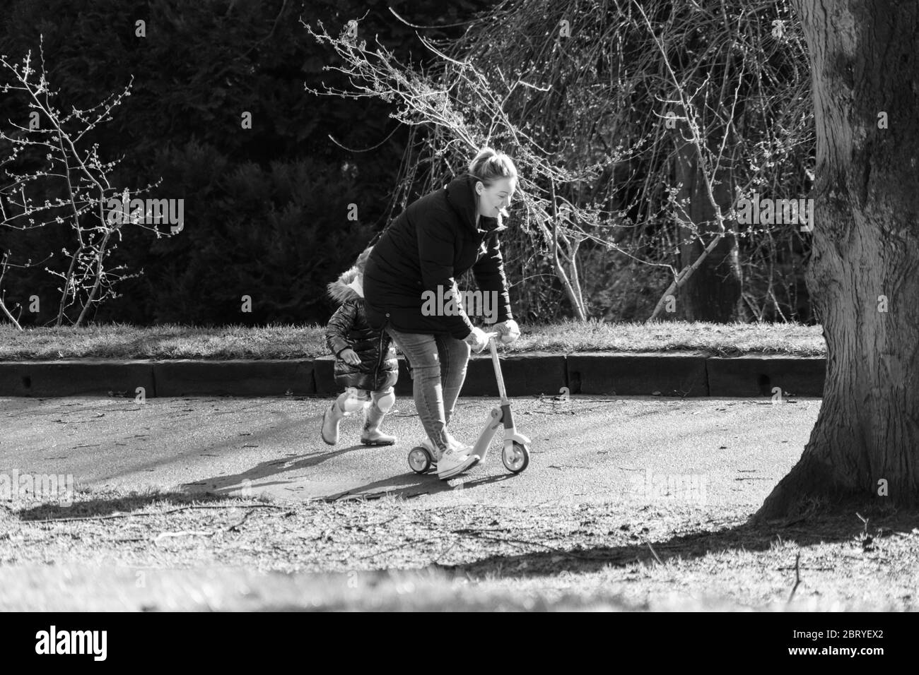 Madre che cavalca giovani figlie scooter in un parco, Valley Gardens, Harrogate, North Yorkshire, Inghilterra, REGNO UNITO. Foto Stock