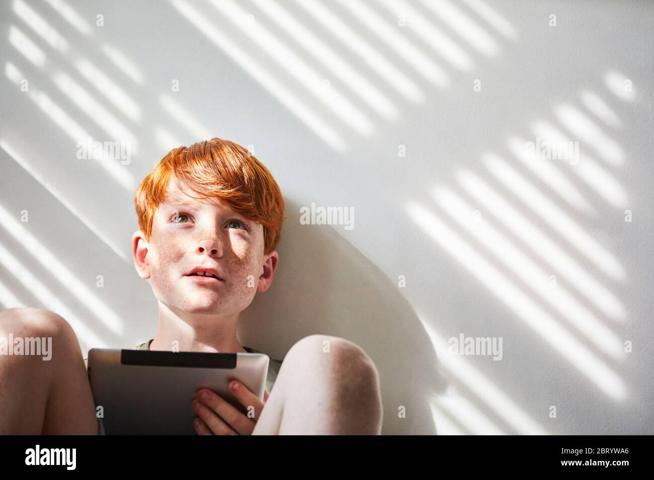 Ragazzo con capelli rossi seduto sul pavimento in una stanza soleggiata, tenendo un tablet digitale. Foto Stock