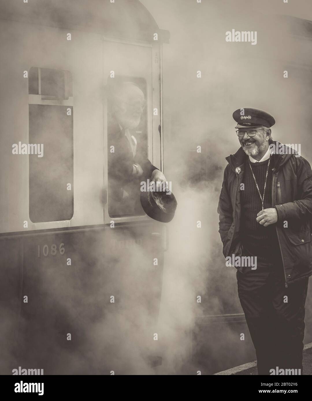 Vista monocromatica di un sorridente lavoratore ferroviario a vapore sulla piattaforma che chiacchiera con l'ispettore dei biglietti attraverso la finestra di lasciare il treno vintage del Regno Unito. Vapore pieno davanti! Foto Stock