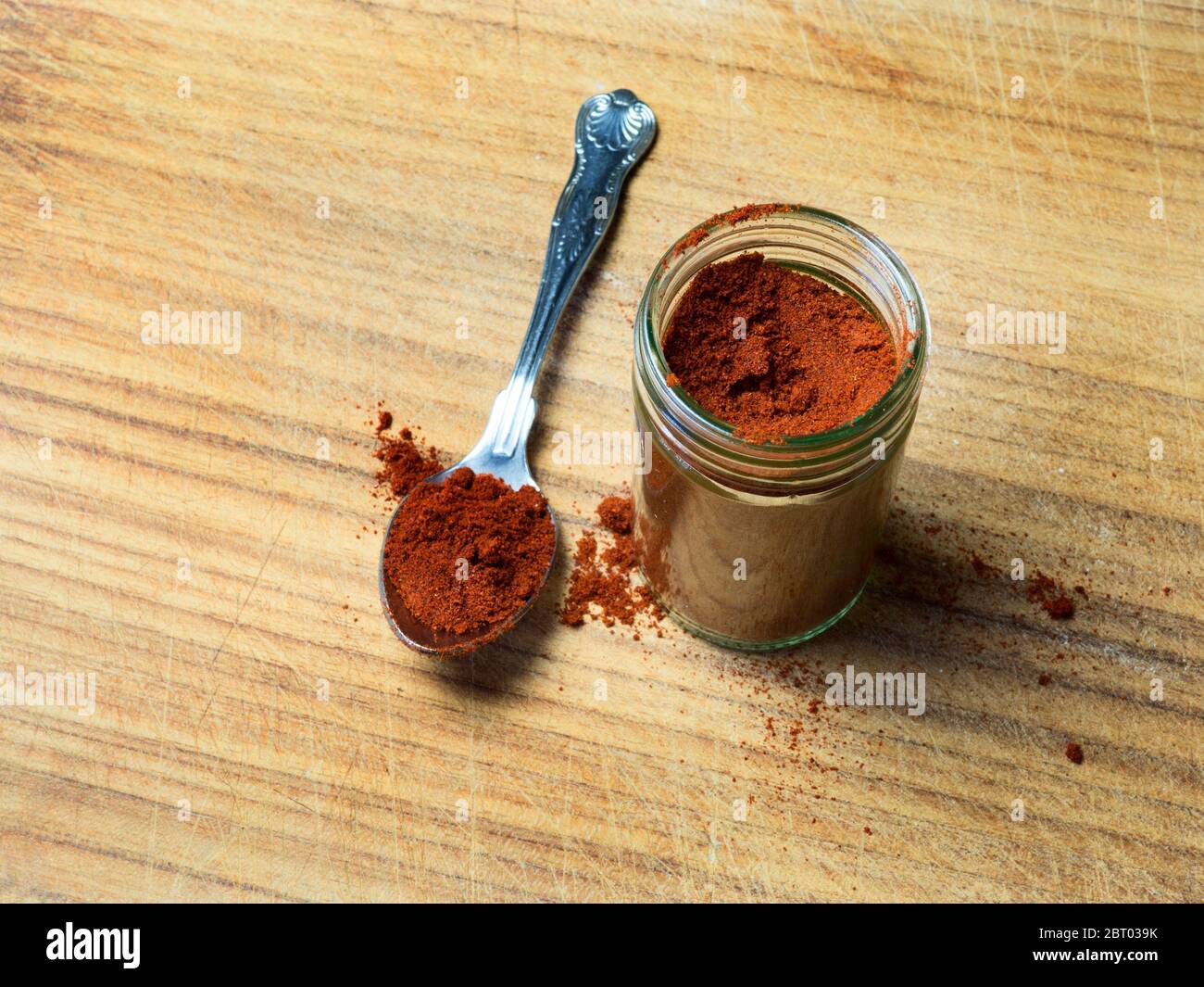 Un vasetto di vetro di peperoncino in polvere caldo con un cucchiaino di peperoncino in polvere caldo su un asse di legno Foto Stock