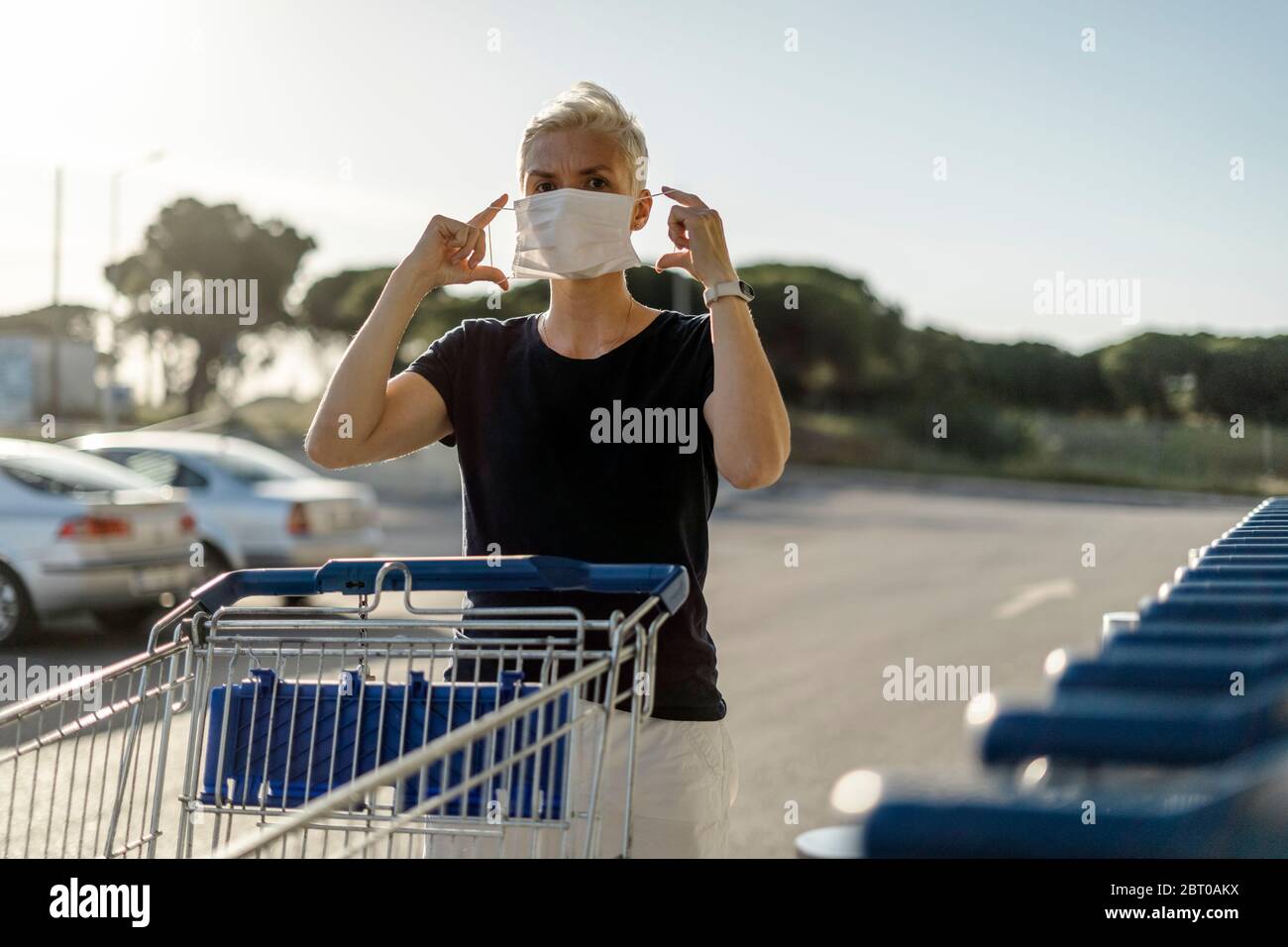 Donna con carrello di shopping che mette la maschera protettiva sul viso appena prima di entrare in un negozio Foto Stock