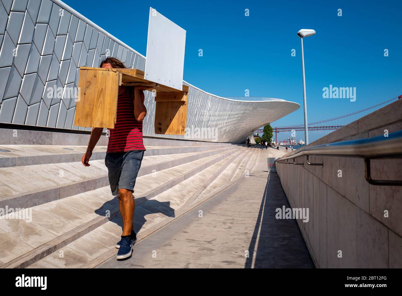 Europa, Portogallo, Lisbona. Un uomo cammina con una panchina sulla spalla di fronte a un edificio a forma di onda a Lisbona. Foto Stock