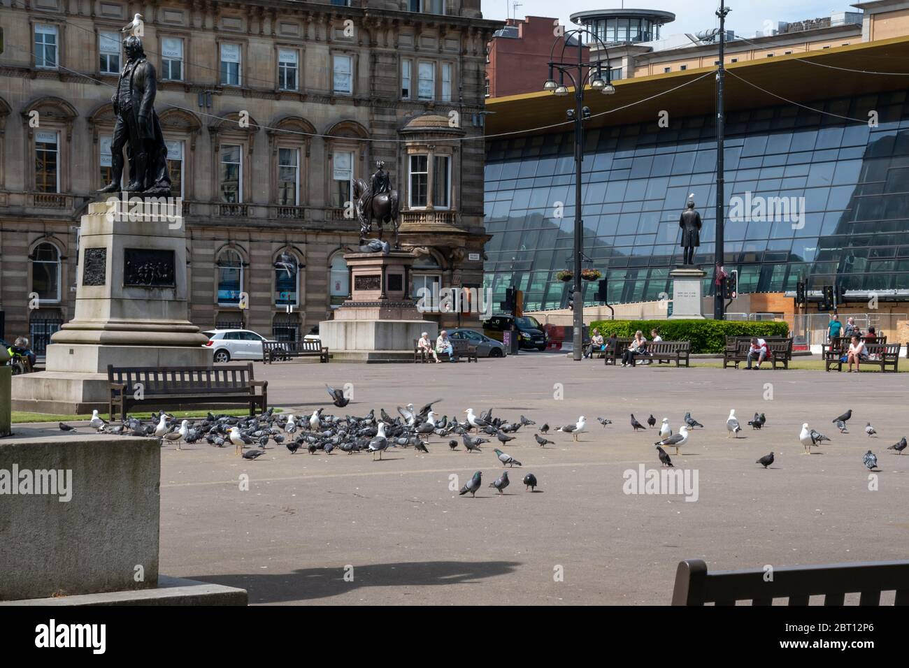 George Square, come la gente social distanza sulle panchine in una calda giornata di sole con la recentemente rinnovata Queen Street Station visibile sullo sfondo. Foto Stock