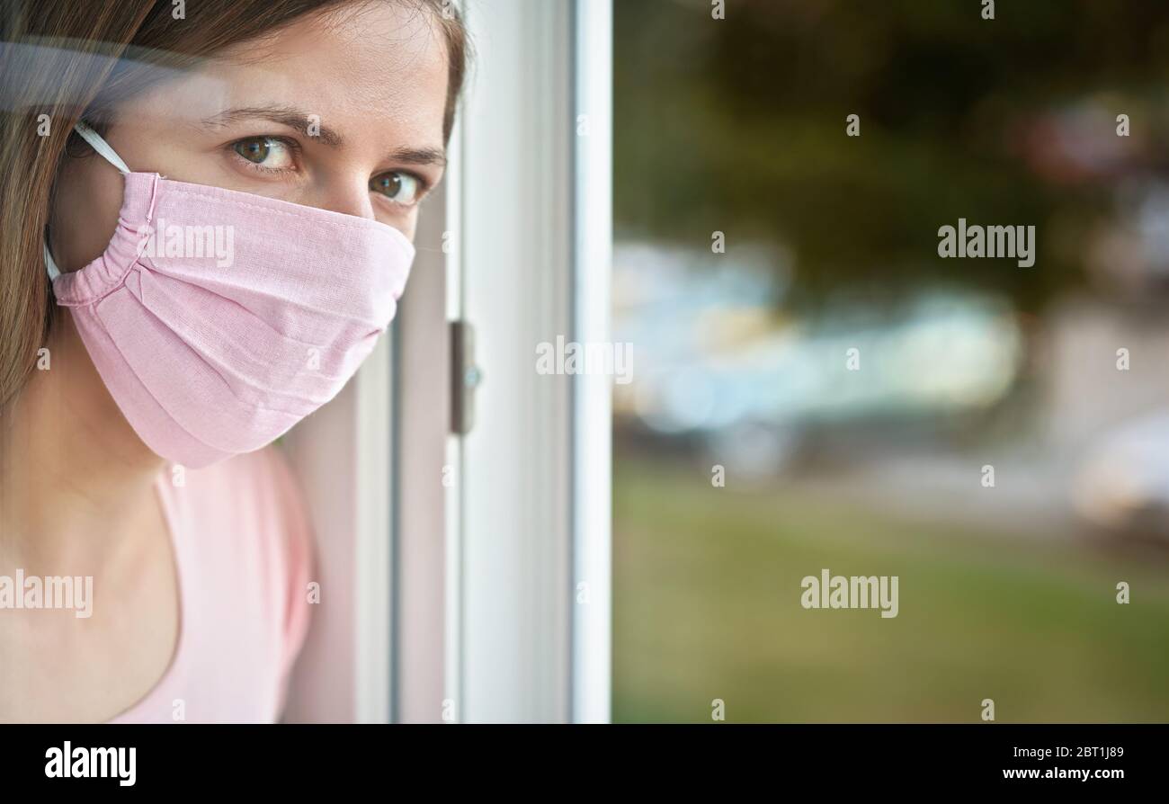 Giovane donna in rosa cotone viso virus maschera in piedi dietro vetro finestra vetro pannello, toccandolo con la mano, guardando triste. Quarantena o soggiorno a casa per essere saf Foto Stock
