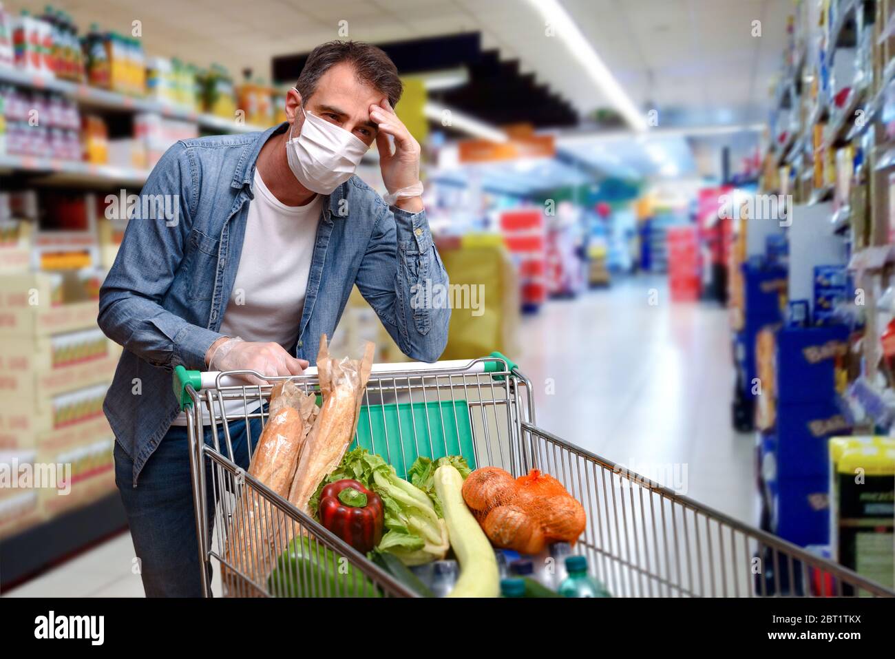 Uomo malato con febbre appoggiato su un carrello pieno di cibo con schermi facciali e guanti in un supermercato Foto Stock