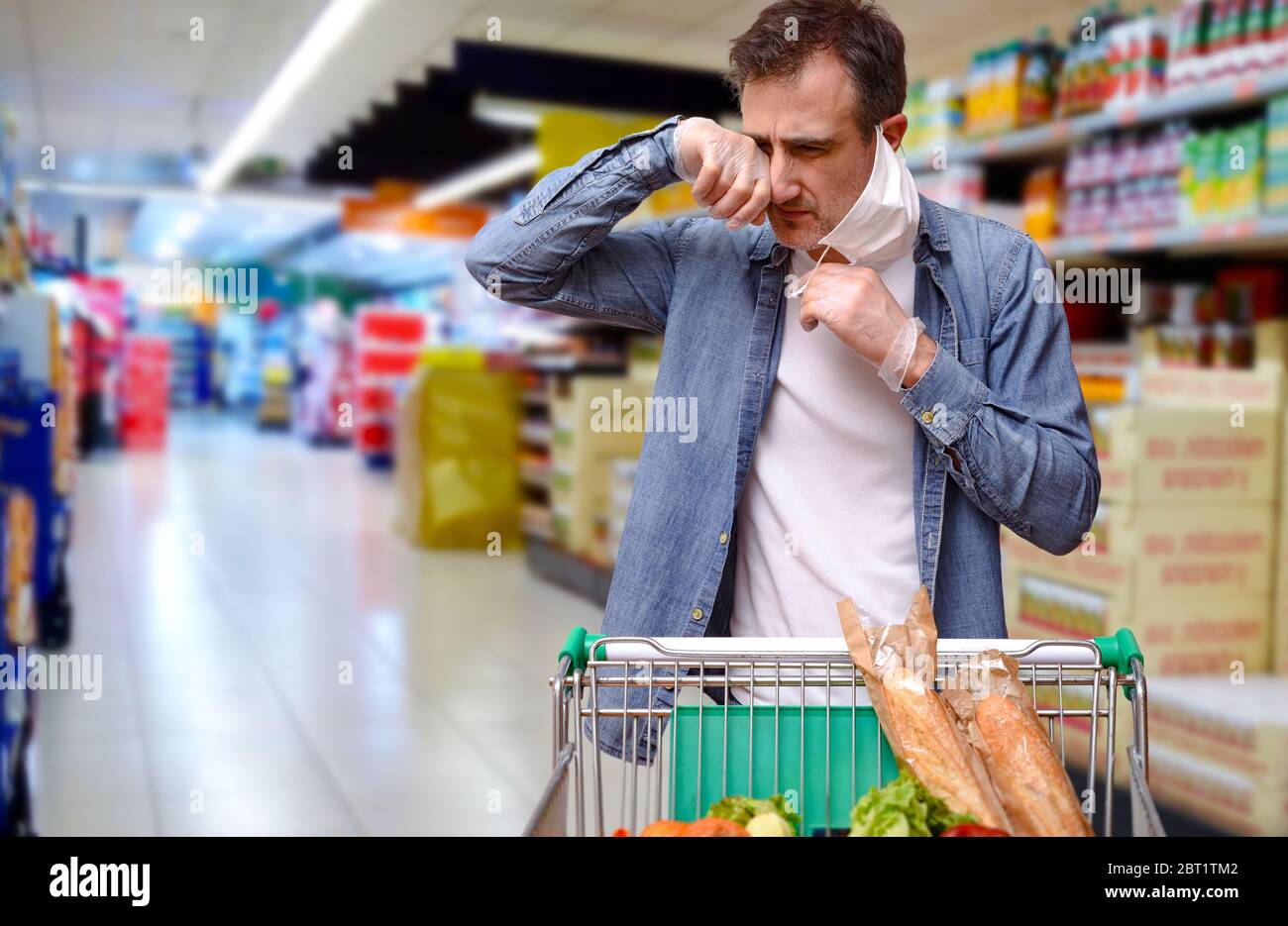 Uomo trascurato che tocca il suo viso sotto la maschera di acquisto in un supermercato con un carrello pieno di cibo Foto Stock