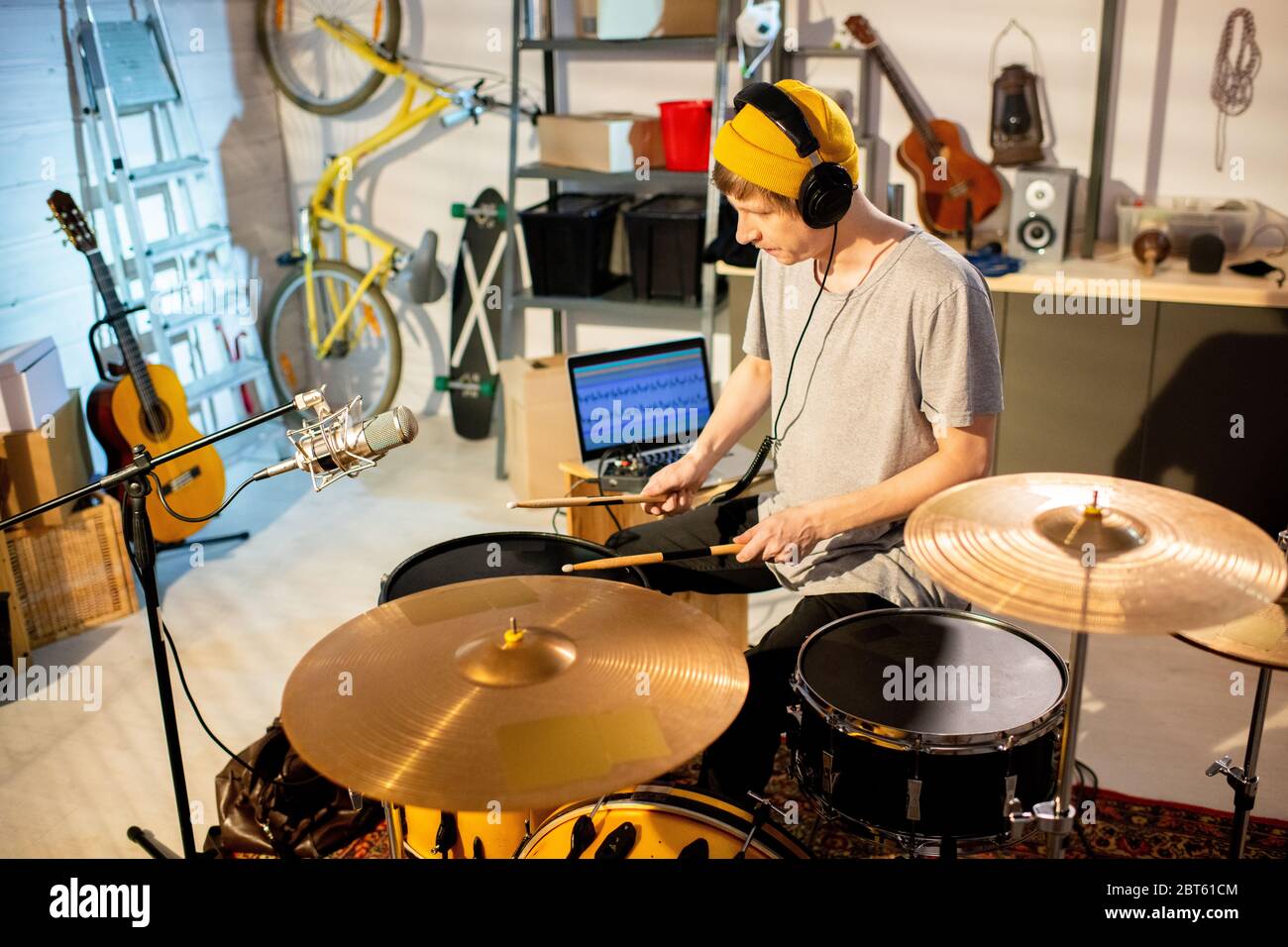 Giovane musicista in t-shirt e bebania colpendo tamburi e cembali con i drumsticks mentre sedendo da batterista nel suo garage o studio Foto Stock