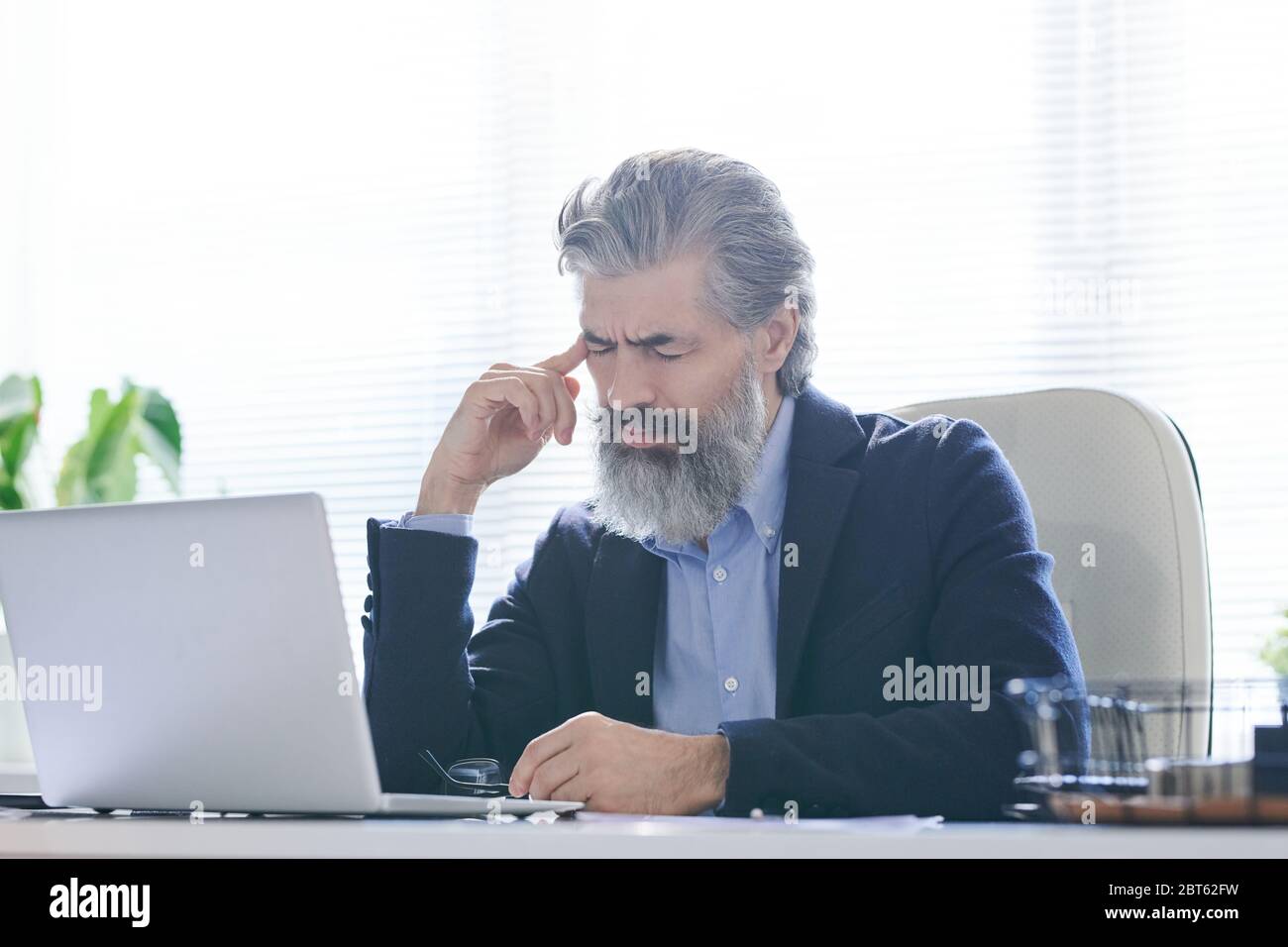 Uomo anziano stanco o pensivo con barba grigia e capelli che toccano il tempio mentre soffre di mal di testa o cercando di ricordare qualcosa Foto Stock